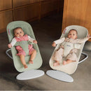 Two infants sitting in Babybjorn Bouncer Begin seats; one has a light green fabric, the other a neutral tone, both feature adjustable harnesses.