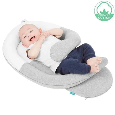 A baby lies comfortably in a grey Babymoov Cloudnest organic cotton lounger, smiling and playing with its hands.