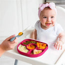 A baby in a high chair smiles while eating from a pink Bbluv Yümi silicone plate with four sections, holding a spoon.