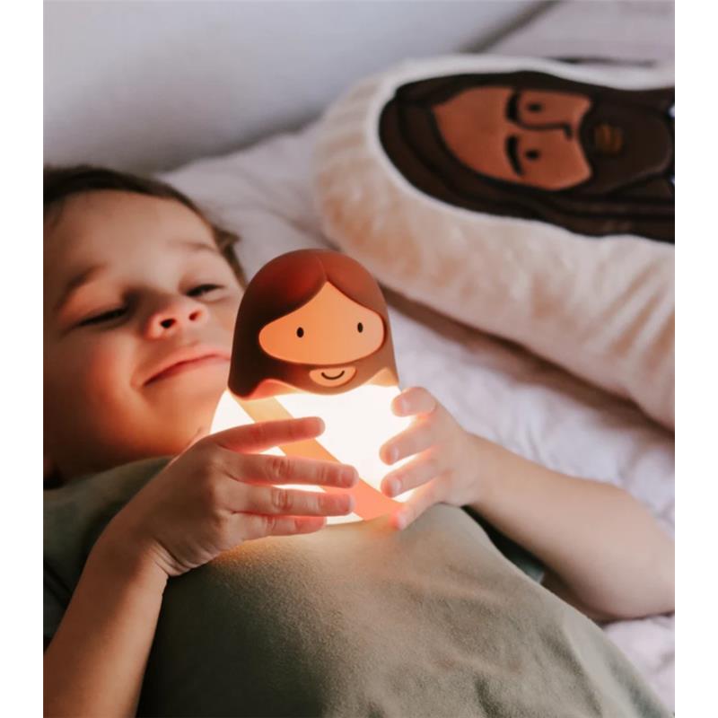 A child smiles while holding a glowing Jesus-themed night light in bed, with a matching pillow featuring a similar design.