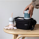 Beaba Stainless Steel Insulated Food Jar 10 oz in sage color being placed into a black bag on a rattan table, with a plush toy nearby.