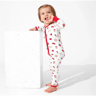 Toddler in a Christmas-themed bamboo footie with gingerbread and Santa designs, playfully leaning on a white pedestal.