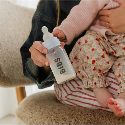 Silicone bottle nipple from Bibs held in a hand, with a baby wearing floral pants visible beside it.