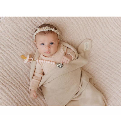 A baby wrapped in a beige blanket lies on a textured background, holding a pacifier and toy.