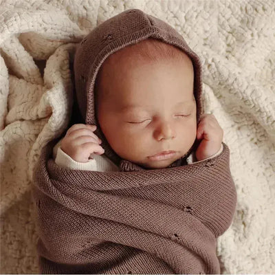 Beige organic cotton blanket and bonnet hat set being used to gently wrap a sleeping newborn.