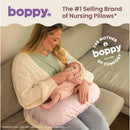 A woman sits on a couch holding a baby and feeding her with a bottle, using a Boppy Nursing Pillow in a blush butterfly print.