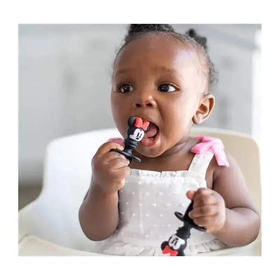 Baby chewing on a black and red silicone utensil shaped like Minnie Mouse, wearing a white dress with pink straps.