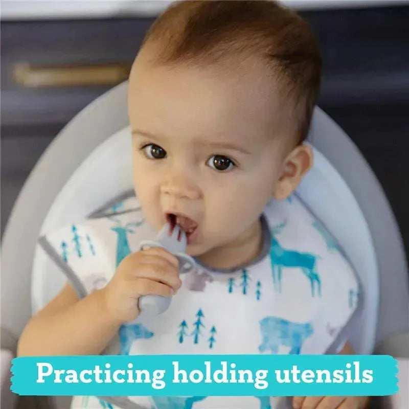 A toddler in a high chair is practicing with a gray silicone Chewtensil, gripping it while bringing it to their mouth.