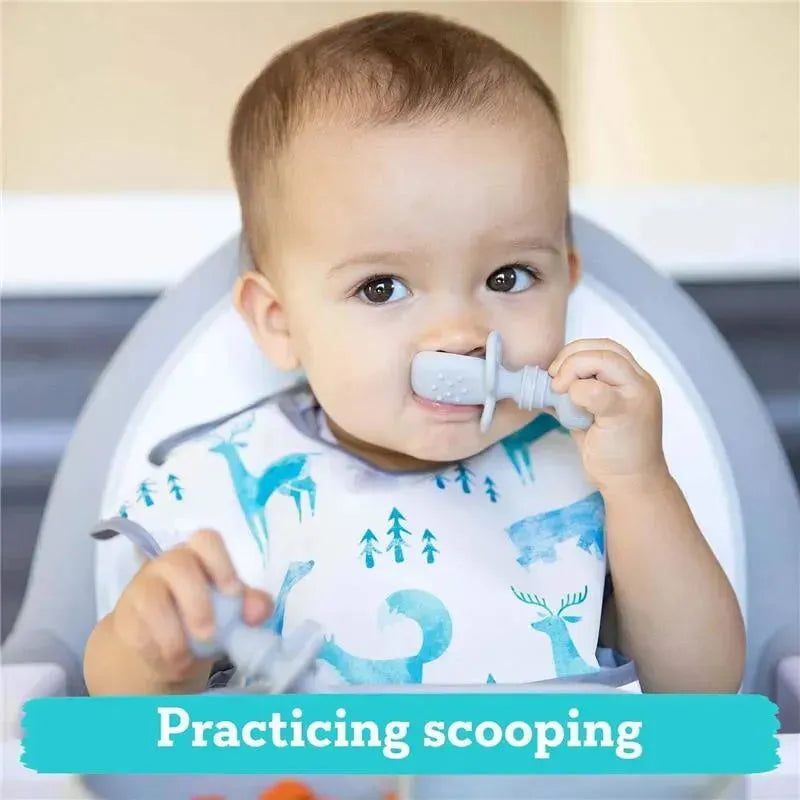 A toddler in a high chair is practicing scooping with a gray Bumkins silicone Chewtensil, smiling while holding it near their mouth.