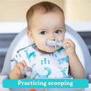 A toddler in a high chair is practicing scooping with a gray Bumkins silicone Chewtensil, smiling while holding it near their mouth.
