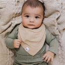 Baby wearing a tan bandana bib, nestled in a soft, textured blanket, with a neutral green outfit and gentle expression.