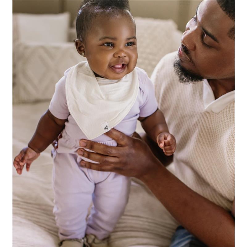 Smiling baby in a light purple outfit wearing a white bandana bib, held by an adult, on a cozy sofa.
