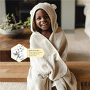 Toddler wearing a beige hooded towel with bear ears, smiling in a cozy indoor setting. Text highlights organic cotton material.