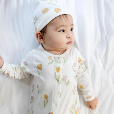 Baby girl wears a Burts Bees jumpsuit and hat set with a floral pattern, lying on a light background.