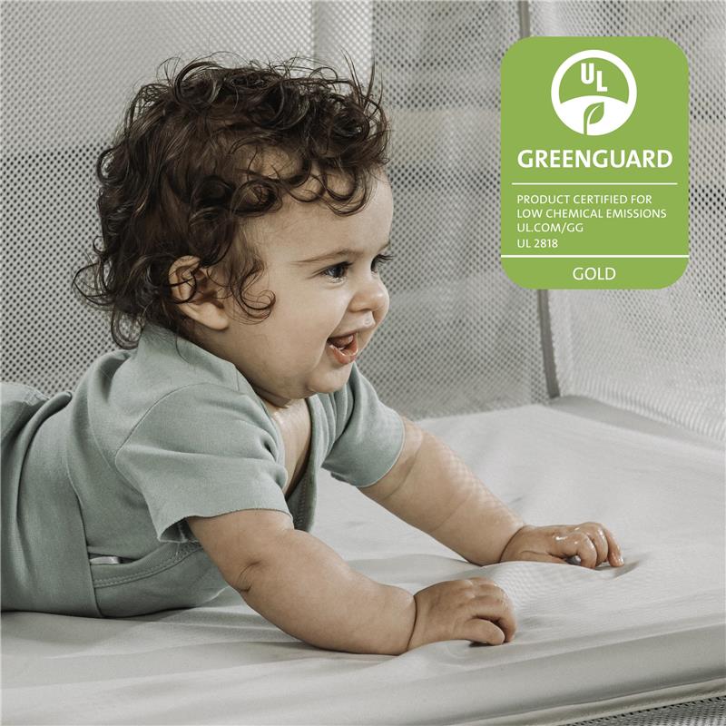A toddler with curly hair smiles while playing on a soft surface inside a portable playard, featuring Greenguard certification.