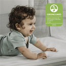 A toddler with curly hair smiles while playing on a soft surface inside a portable playard, featuring Greenguard certification.