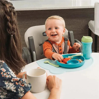 Chicco Polly highchair in taupe with a smiling toddler eating from a blue plate, holding a sippy cup, at a table.