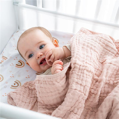 Baby smiling while wrapped in a blush pink Comfy Cubs muslin blanket in a crib with patterned bedding.