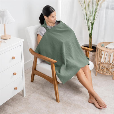 A woman sitting in a chair uses a fern-colored muslin nursing cover, providing privacy while nursing.