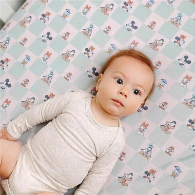 A baby lies on a pastel crib sheet featuring Disney characters Mickey Mouse and friends in a playful pattern.