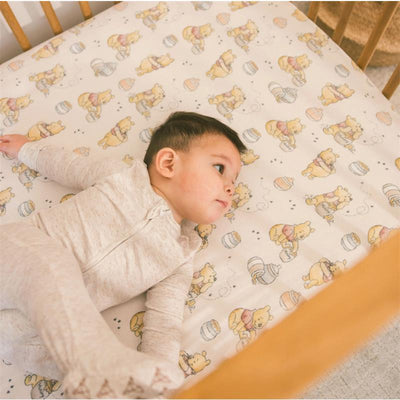 Premium crib sheet featuring a whimsical Winnie the Pooh design with a baby lying on it in a crib.