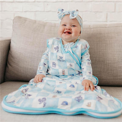 Baby wearing a blue-themed Copper Pearl Monsters Inc Medley 0.5 TOG sleep bag, smiling and seated on a couch.
