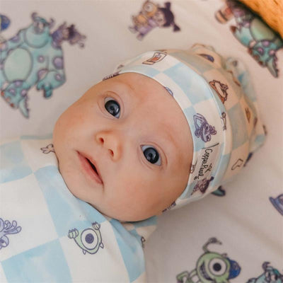 Infant wearing a Copper Pearl Monsters Inc Medley Top Knot Hat, featuring a blue checked design with cartoon monsters.