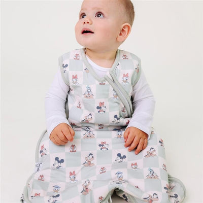 A toddler in a Mickey Mouse-themed sleep bag sits on the floor, looking up with curiosity.