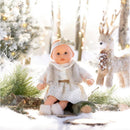 Bébé Calin Margaux doll in a winter scene, wearing a white dress with a fur vest and a bow, surrounded by snow and greenery.