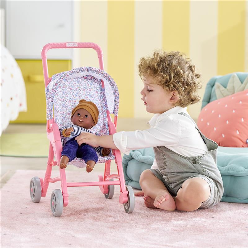 Child playing with a pink floral pushchair, holding a 30 cm doll dressed in a striped outfit and brown hat.