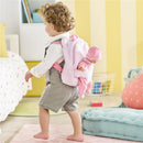 Child wearing a pink plush backpack carrier with a baby doll inside, walking in a brightly colored room.