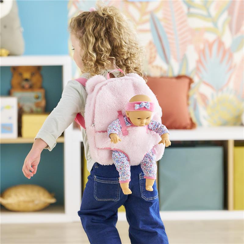 Plush pink backpack with a baby doll in a floral outfit attached, worn by a child in a playful indoor setting.
