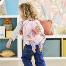 Plush pink backpack with a baby doll in a floral outfit attached, worn by a child in a playful indoor setting.