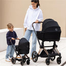 A woman in a white hoodie pushes a black pram while a young child happily holds a smaller doll stroller.
