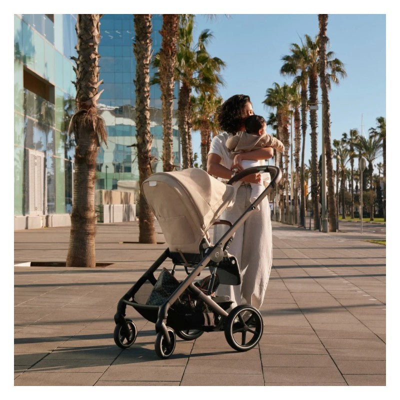 Cybex Balios S Lux stroller in Stone Grey parked on a sunny walkway, with a mother holding a baby nearby.
