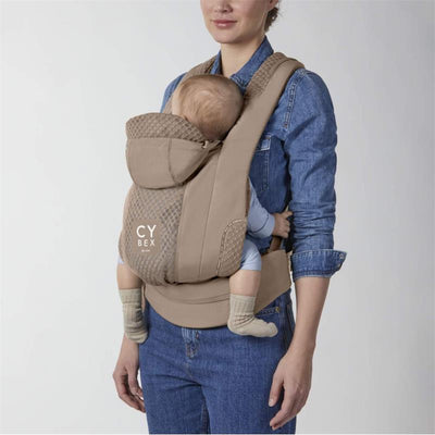 Cybex Coya Carrier in Cozy Beige, worn by a woman with a baby securely seated, featuring a hood and textured design.