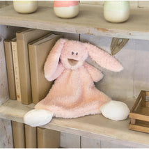 Soft pink bunny rattle blankie with floppy ears and a smiling face, sitting on a wooden shelf near books and decorative items.