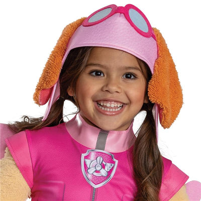 Toddler girl wearing a pink Paw Patrol Skye costume with goggles and dog-ear headband, smiling joyfully.