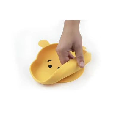 Yellow silicone grip dish shaped like Winnie the Pooh, being held with one hand, showing its flexible design.