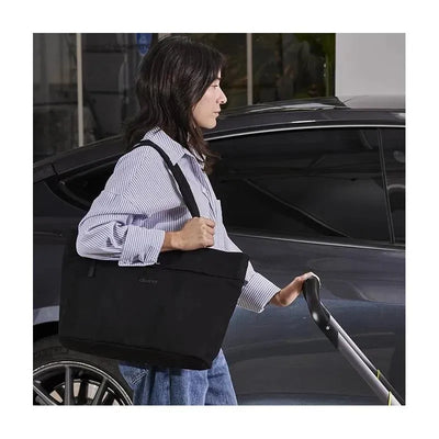 A woman carrying a black Doona Essentials Diaper Tote Bag while holding the handle of a stroller near a car.