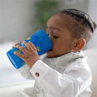 Toddler drinking from a blue Dr. Brown Cheers360 training sippy cup with green leaf designs while wearing a white shirt.