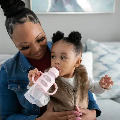 Light pink Dr. Brown sippy spout bottle with silicone handles, held by a toddler, smiling while drinking.