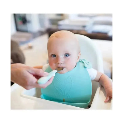 A baby in a high chair wearing a teal bib is being fed with a soft grip spoon by an adult.