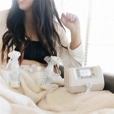 Black hands-free pumping bra with breast pumps attached, worn by a woman sitting and smiling, near a breast pump device displaying settings.