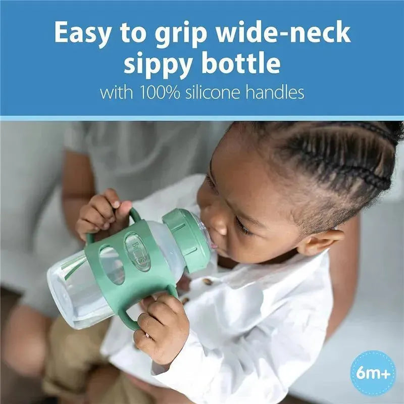 Wide-neck sippy spout bottle with green silicone handles being used by a child, suitable for ages 6 months and up.