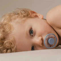 A young child with curly blonde hair lies on a soft surface, sucking on a blue pacifier.