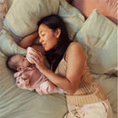 A mother gently feeds her baby with a bottle while lying on soft pillows, both looking calm and content.