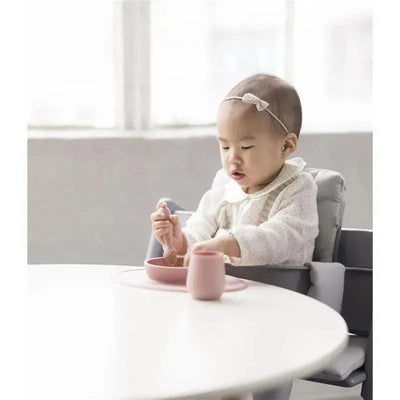 Baby using the Ezpz First Foods Set in blush color, sitting at a table with a bowl and cup, engaged in eating.
