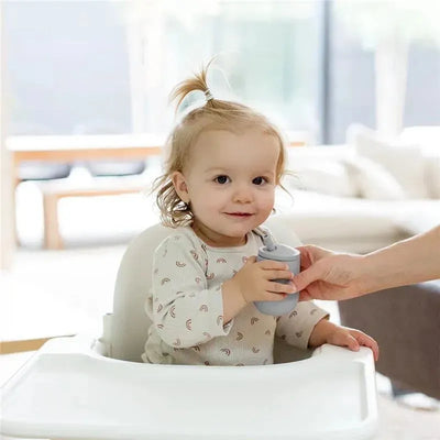 Toddler in a high chair holds a gray Ezpz Mini Cup & Straw Training System, smiling at an adult handing it to them.