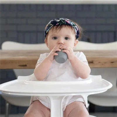 Baby sitting in a high chair, holding a gray Ezpz Tiny Cup, wearing a floral headband and a white outfit.